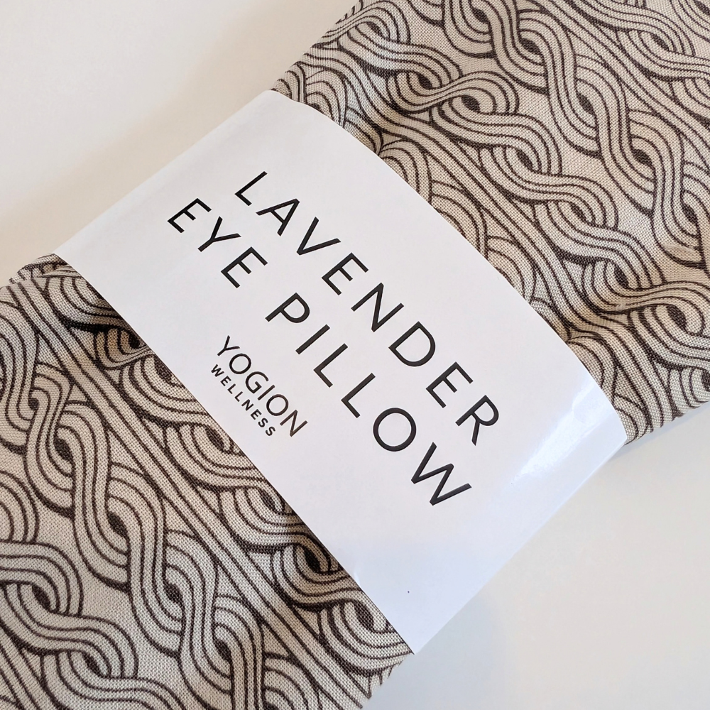 A folded eye pillow with a beige and brown wavy pattern, wrapped in a white paper band that reads 'LAVENDER EYE PILLOW' and 'YOGLON WELLNESS'.