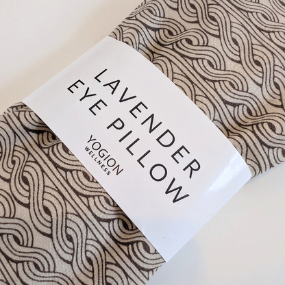 A folded eye pillow with a beige and brown wavy pattern, wrapped in a white paper band that reads 'LAVENDER EYE PILLOW' and 'YOGLON WELLNESS'.