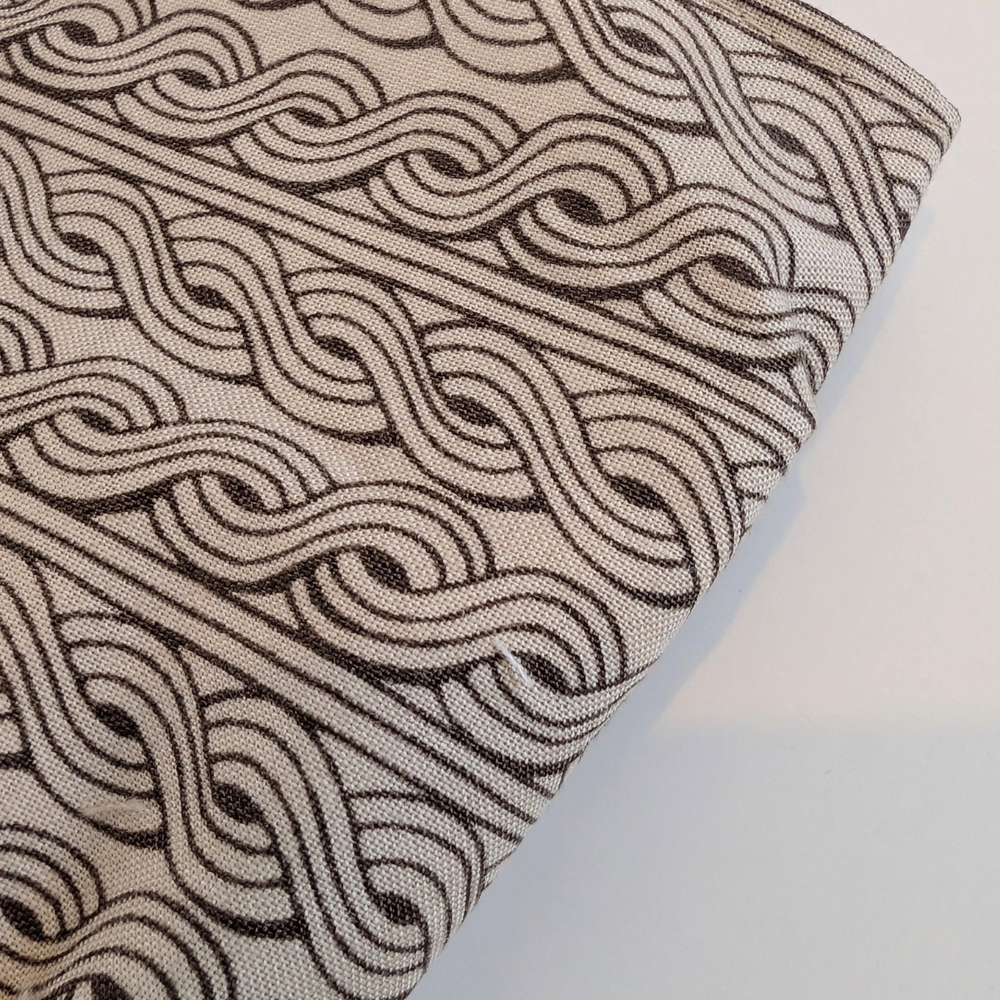 A close-up of a woven fabric with a repeating black and beige wavy interlocking pattern, folded diagonally on a light gray background.