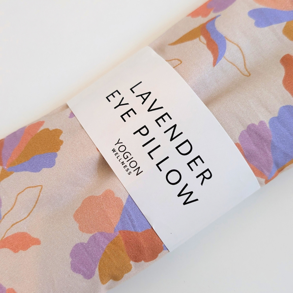 A rolled fabric eye pillow with a lavender floral pattern in purple, orange, and blue on a light beige background, wrapped in a white paper band that reads 'LAVENDER EYE PILLOW' and shows the brand 'YOGION WELLNESS'.