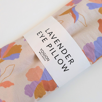 A rolled fabric eye pillow with a lavender floral pattern in purple, orange, and blue on a light beige background, wrapped in a white paper band that reads 'LAVENDER EYE PILLOW' and shows the brand 'YOGION WELLNESS'.