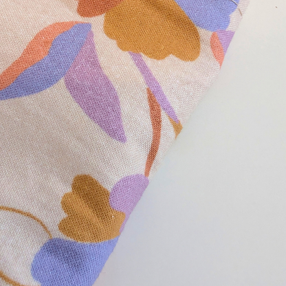 A close-up of a woven fabric with a retro floral pattern in purple, orange, and mustard yellow on a cream background, shown against a plain white surface.
