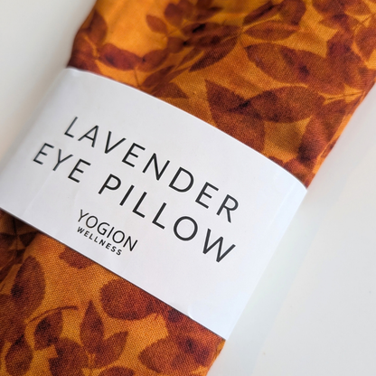 An eye pillow with a brown and orange leaf pattern, wrapped in a white paper band that reads 'LAVENDER EYE PILLOW' by 'YOGION WELLNESS'.