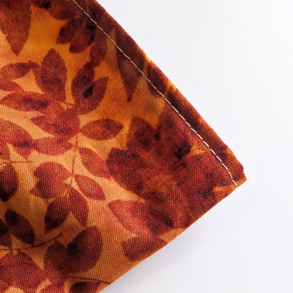 A close-up of a folded textile corner showing a warm orange and brown leaf pattern with visible white stitching on a plain white background.