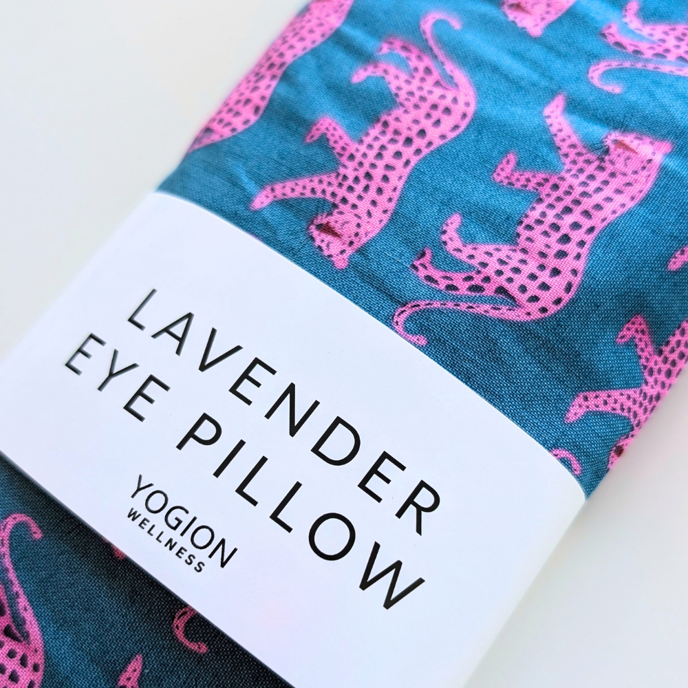 A folded eye pillow with a teal blue background and pink spotted animal pattern, wrapped with a white paper band labeled 'LAVENDER EYE PILLOW' and 'YOGION WELLNESS'.