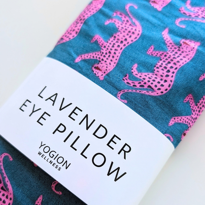 A folded eye pillow with a teal blue background and pink spotted animal pattern, wrapped with a white paper band labeled 'LAVENDER EYE PILLOW' and 'YOGION WELLNESS'.