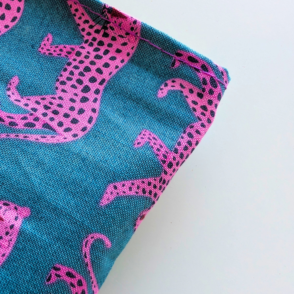 A close-up of a woven textile with a bright teal background and bold pink animal silhouettes outlined in black dots, shown folded on a plain white surface.