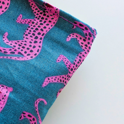 A close-up of a woven textile with a bright teal background and bold pink animal silhouettes outlined in black dots, shown folded on a plain white surface.
