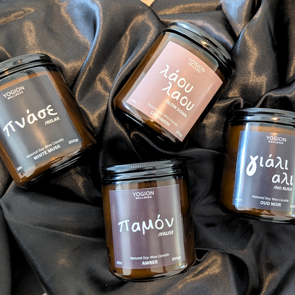 Four amber glass soy wax candles by YOGION WELLNESS arranged on draped black satin fabric. The labels show different scents in Greek: ΦηδιΩΣ (White Musk), Χδθδι δθδι (Oud Corom), Φηδι δθδι (Oud Noir), and Φηδι δθδι (Amber).