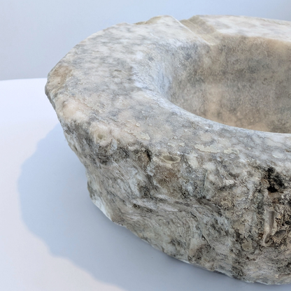 A close-up of a rustic stone basin or sink with a heavily textured, weathered surface in shades of gray, beige, and white, set against a plain light gray background.