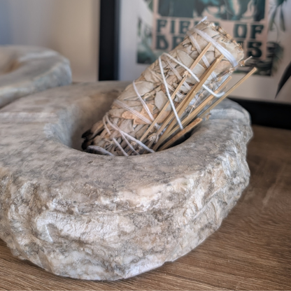 A rustic stone smudge holder containing a bundle of dried sage leaves wrapped in white string, placed on a wooden surface with a blurred framed print in the background.
