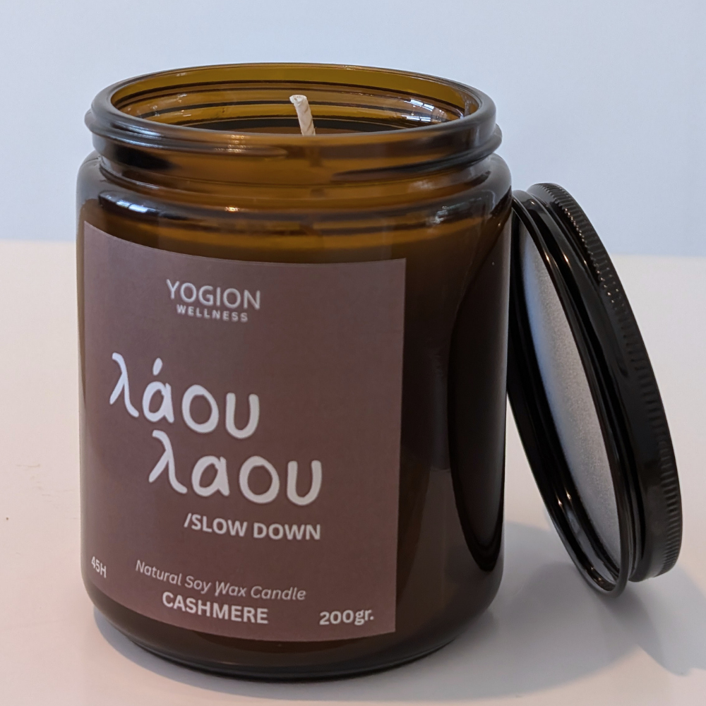 An amber glass jar candle with its lid leaning against it on a white surface. The label reads 'YOGION WELLNESS', 'láou láou / SLOW DOWN', and 'CASHMERE'.