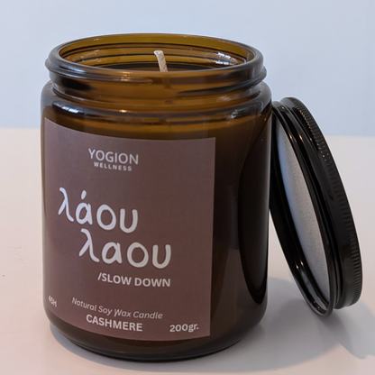 An amber glass jar candle with its lid leaning against it on a white surface. The label reads 'YOGION WELLNESS', 'láou láou / SLOW DOWN', and 'CASHMERE'.