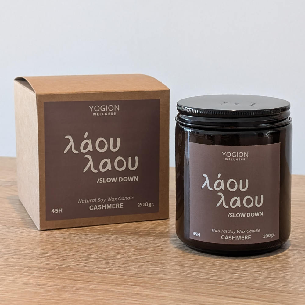 A YOGION WELLNESS soy wax candle in a dark amber glass jar with a black lid, shown next to its brown cardboard box on a light wooden surface against a neutral light gray background.