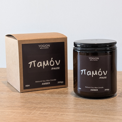 A YOGION WELLNESS παμόν amber soy wax candle shown next to its kraft paper box on a light wooden tabletop against a neutral light gray background.