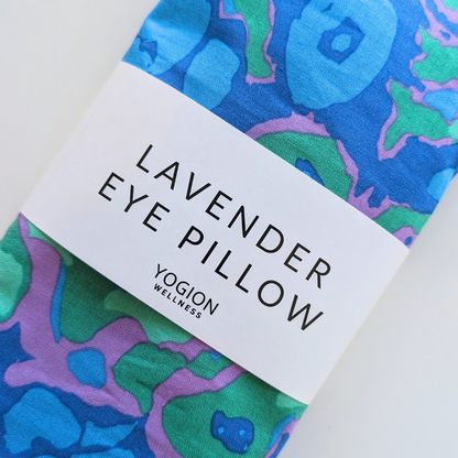 A close-up of a folded eye pillow sham with a colorful abstract watercolor pattern in blue, green, and purple, wrapped with a white paper band labeled 'LAVENDER EYE PILLOW' and 'YOGION WELLNESS'.