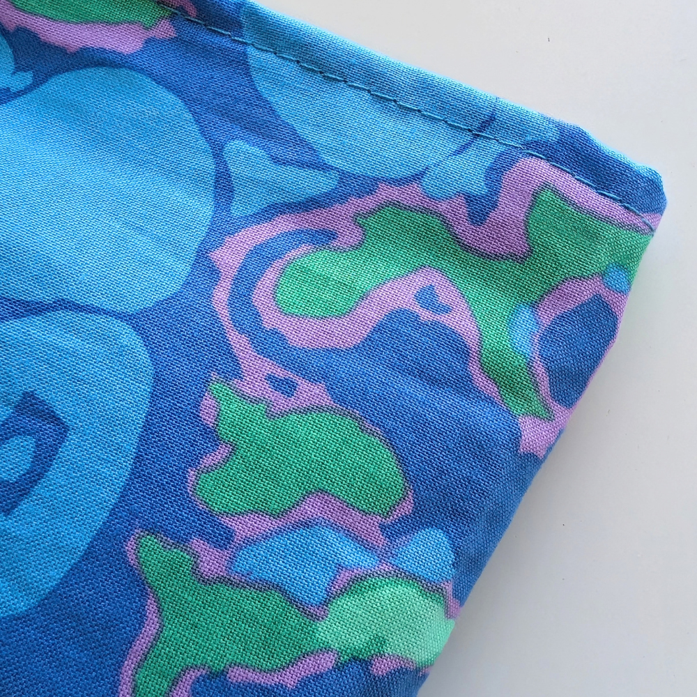 A close-up of a folded piece of fabric with a retro abstract pattern in blue, green, and pink, showing the woven texture and neat hem on a white background.