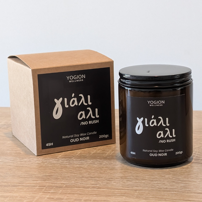 A YOGION WELLNESS candle in an amber glass jar with a black lid, shown next to its brown kraft paper box on a light wooden surface against a white background. yiali ali 