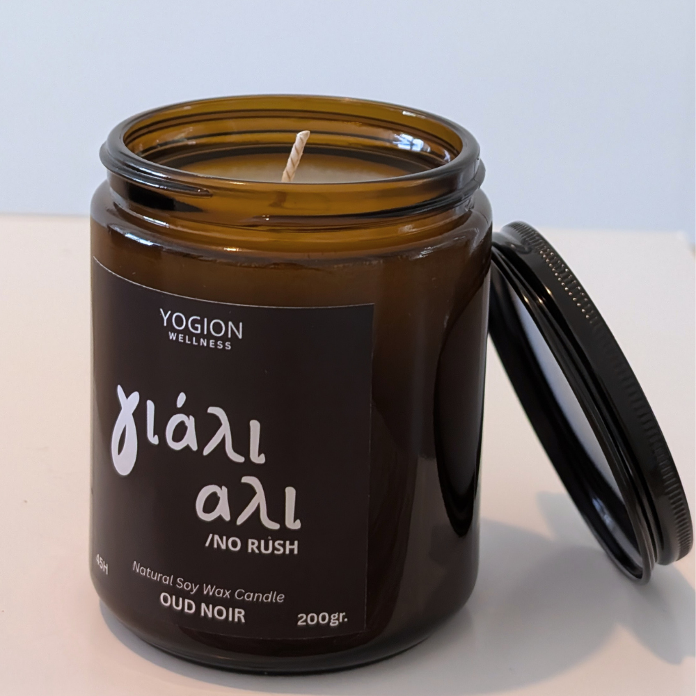 An amber glass jar candle with its lid leaning against it on a white surface. The label reads 'YOGION WELLNESS' and 'Oud Noir / No Rush'.