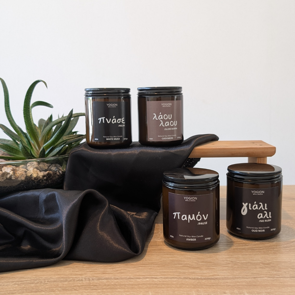 Four dark amber glass candles with black lids and white Greek labels are arranged on a wooden surface with a draped black fabric and a small plant in the background. Visible labels include yogion wellness scents such as Πνάσε, Λάου λαου, Παμόν, and γιαλι Αλι.